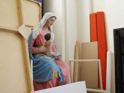Una statua della Vergine col bambino in abiti rosa e azzurro, giace invece silenziosa dietro tele e cornici di legno 